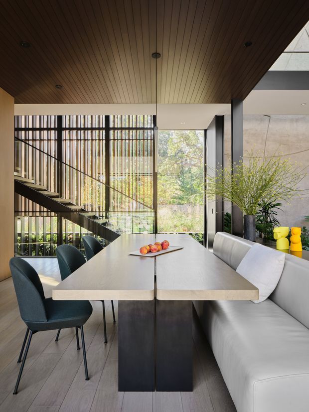 A contemporary dining area in a dramatic open-plan interior, featuring a long pale stone dining table with dark pedestal bases, upholstered dining chairs, and a built-in white banquette. A sculptural open-riser staircase with glass balustrade and black metal framing rises beside tall glazed openings with vertical screening. The room has a warm timber slatted ceiling, light timber flooring, dark structural elements, and large indoor plants, creating a refined, light-filled atmosphere.
