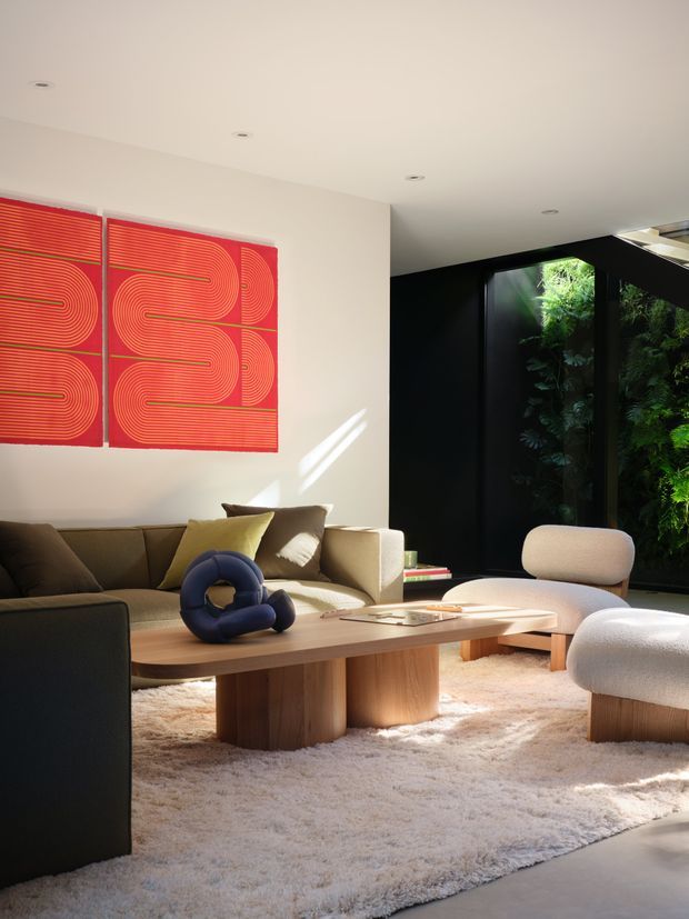 A contemporary living room with a low, sculptural timber coffee table, neutral upholstered seating, a dark sofa, and a large cream shag rug. A bold red geometric artwork hangs on the light wall, while a black-framed glazed opening reveals lush greenery and brings strong natural light into the room. The space has recessed ceiling lights and a refined, minimalist furniture arrangement.