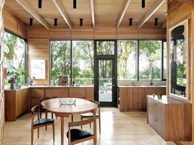 Warm, timber-lined open-plan kitchen and dining area with extensive black-framed glazing overlooking greenery. The kitchen features wood cabinetry, pale timber flooring, a central dining table with mid-century-style chairs, exposed white ceiling beams, and integrated cabinetry along the right wall. Natural daylight creates an inviting connection to the outdoors.