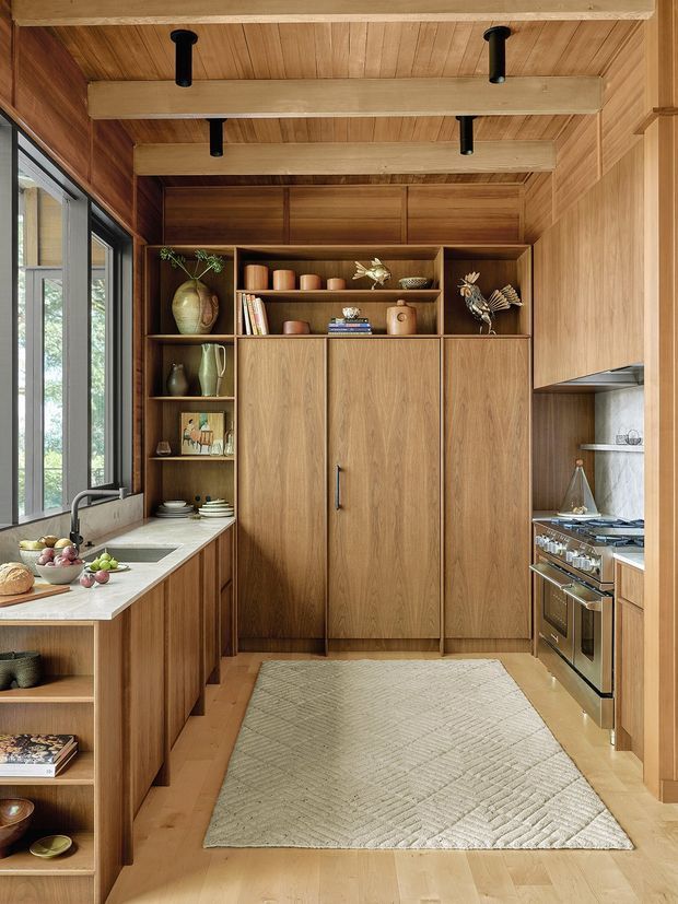 Warm, compact kitchen with extensive natural timber cabinetry and wall panelling, exposed timber ceiling beams, open shelving, a stone or light solid-surface benchtop, integrated sink, and stainless-steel cooking appliances. Tall black-framed glazing brings in daylight along the left side. The space has a refined, natural, bespoke built-in character with earthy decorative accents and a large neutral textured rug.