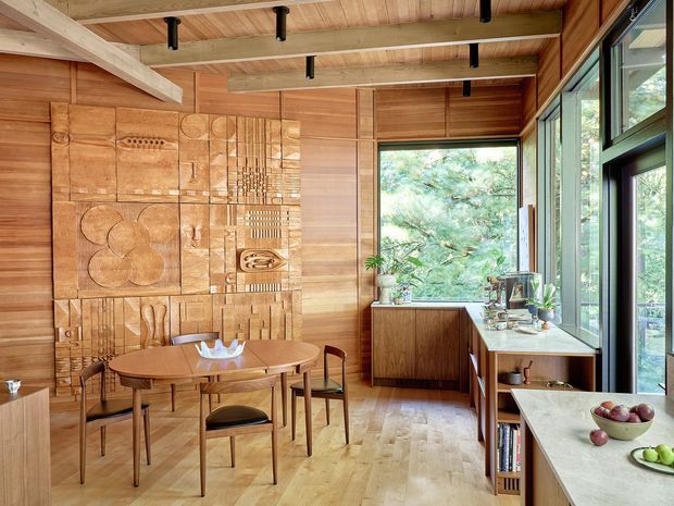 Warm timber-lined dining and kitchen space with a mid-century-inspired round dining table and chairs, a sculptural carved timber feature wall, exposed timber ceiling beams, and extensive glazing overlooking greenery. A compact timber kitchen or serving area with stone benchtops occupies the right side, creating an open, light-filled interior.