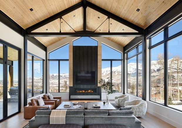 A spacious contemporary living room with a dramatic vaulted timber ceiling and exposed black steel trusses. Floor-to-ceiling glazing wraps the room, offering expansive views of snow-covered mountain ranges. A central dark feature wall incorporates a television and linear fireplace, while layered sofas, armchairs, and a coffee table create a refined seating area.