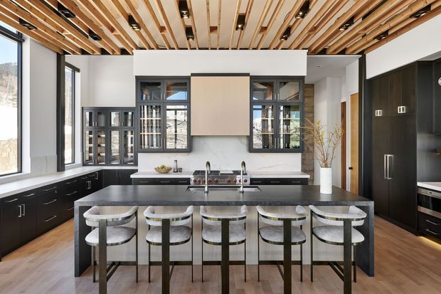 A spacious contemporary kitchen with a large dark stone island and five bar stools, matte black cabinetry, integrated appliances, expansive black-framed windows, and a dramatic slatted timber ceiling with recessed lighting. Light timber flooring and pale walls balance the dark joinery, while a central timber-toned rangehood surround forms a focal point.