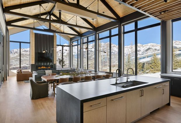 A spacious open-plan kitchen and living area with a large central island, integrated sink and dark benchtop, light timber cabinetry, and extensive black-framed floor-to-ceiling glazing. The living room includes a dining table, sofas, and a fireplace wall, while exposed dark structural beams and timber ceiling lining create a warm mountain-home interior. Snow-covered mountains are visible beyond the glazing.