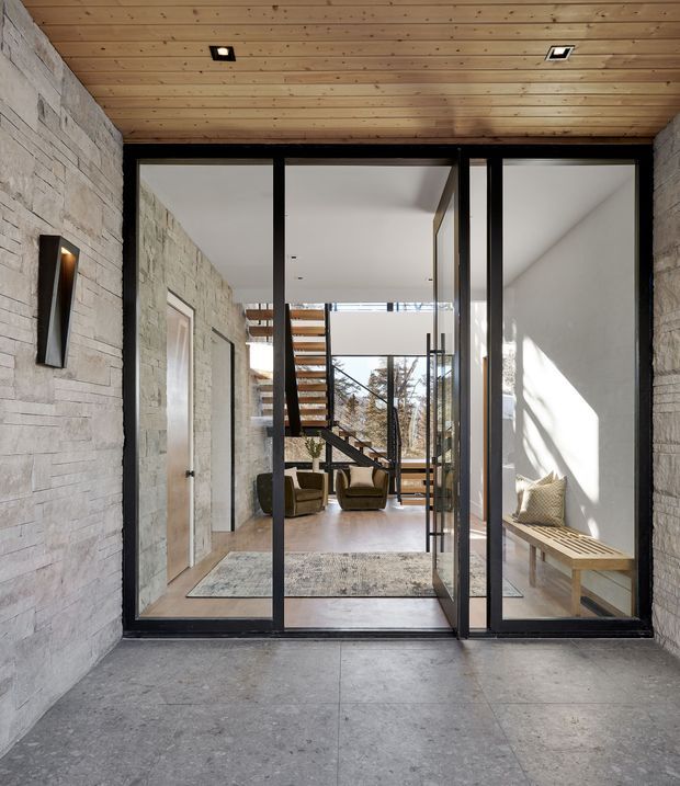 A modern residential entryway viewed from outside through a large black-framed glazed entrance. The entry has pale stacked-stone walls, a warm timber-lined ceiling, and a grey stone or concrete floor. Inside, a timber staircase with open risers leads to an upper level, with lounge chairs, a rug, a bench, and abundant natural light visible beyond the glass.