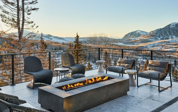 An elevated outdoor terrace overlooking a snow-covered alpine mountain landscape. The terrace has dark tiled or stone paving, black metal balustrades, woven lounge chairs, small side tables, and a large rectangular built-in concrete fire table with visible flames. The scene has a refined contemporary outdoor-lounge character and warm evening light.