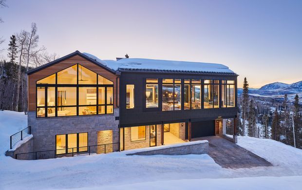 A contemporary two-storey mountain house set in a snowy, wooded landscape. The building has extensive floor-to-ceiling glazing, a steeply pitched gabled volume on the left, and a long glazed rectangular volume on the right. Dark exterior cladding contrasts with grey masonry, warm timber-toned interiors, and snow-covered terrain. A mountain ridge is visible in the background.