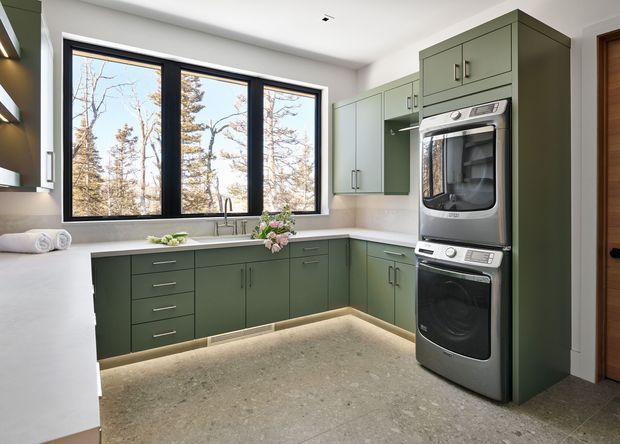 A bright, contemporary kitchen with dark sage-green built-in cabinetry, pale stone benchtops, integrated stainless-steel double oven, and a large black-framed window overlooking trees. Under-cabinet lighting, a central sink, and streamlined hardware create a clean, refined fit-out.