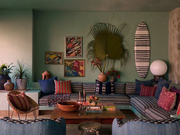 Eclectic living room with a low, built-in striped sectional sofa wrapping around the space, colourful patterned cushions, a woven lounge chair, wooden coffee table, tropical plants, framed artwork, a surfboard-shaped wall accent and a spherical floor lamp. The room has muted green walls, a textured stone or plaster column, and a warm, richly layered palette.