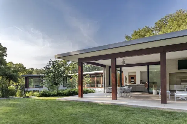 Exterior view of a low-slung modern home opening onto a covered outdoor living and dining terrace. The flat-roofed pavilion has dark timber posts and beams, pale rendered or masonry walls, broad glazing, and a seamless connection to a landscaped lawn and mature trees. A living area is visible through the open terrace, with a separate pavilion or wing in the background.