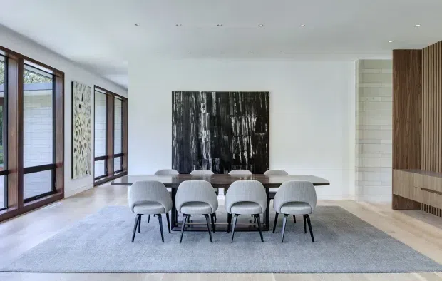 Contemporary dining room with a long dark wood dining table, upholstered grey dining chairs, and a large grey area rug. Full-height dark-framed windows line the left side, while a white tiled fireplace surround and integrated dark timber feature joinery define the right wall. A large monochrome artwork provides a dramatic focal point.