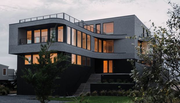 A contemporary multi-storey residence with a sculptural, curving facade, dark horizontal cladding, extensive warm-lit glazing, and a rooftop terrace with slim metal balustrades. The house is viewed externally in a suburban landscaped setting at dusk, with stepped entry stairs and layered balconies/terraces.