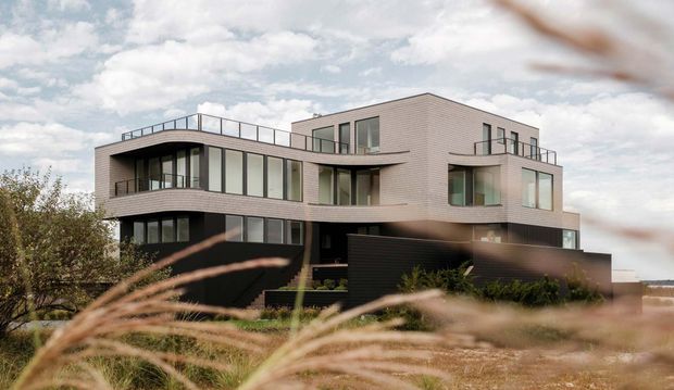 Exterior architectural photograph of a contemporary multi-storey residence with a sculptural curved corner, extensive floor-to-ceiling glazing, pale brick or masonry cladding, dark lower-level walls, and rooftop terraces with slim metal balustrades. Ornamental grasses partially frame the foreground.