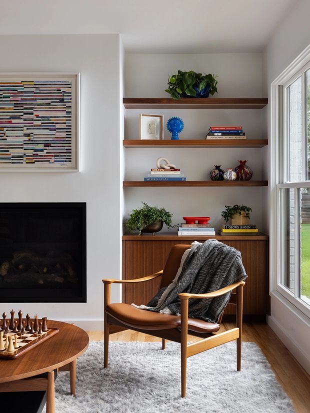 A stylish living room corner with a mid-century-inspired leather lounge chair draped in a grey throw, positioned beside a built-in walnut-toned shelving and cabinetry unit. The shelves display books, plants, ceramics, and decorative objects. A black fireplace and abstract artwork sit to the left, while a large white-framed window brings in natural light from the right. A round timber coffee table with a chess set and a soft grey rug complete the warm, curated interior.
