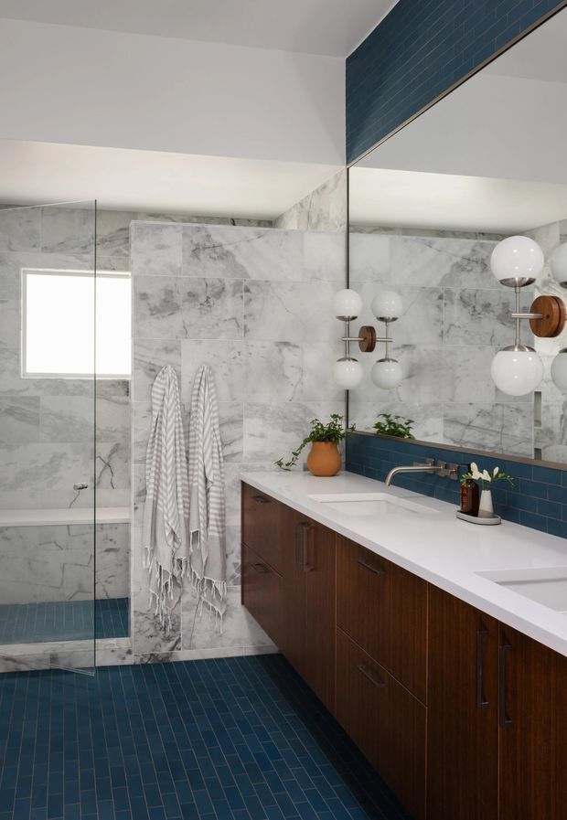 A refined bathroom with a long double vanity, white countertop and integrated sinks, dark wood cabinetry, blue rectangular wall and floor tiles, and large grey-white marble-look wall tiles. A frameless glass shower sits beside the vanity, with a small bathtub visible behind it. The space includes a large mirror, globe wall sconces, potted greenery, and a bright window.