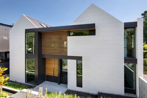 Modern two-storey residential facade with crisp white masonry cladding, steep gabled roof forms, dark charcoal framing, tall vertical glazing, and a recessed entry. Warm timber slatted screening and a timber front door add contrast, while landscaped planting borders the paved approach.
