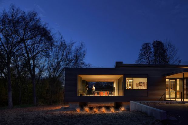 A low, single-storey modern house photographed at dusk, set among mature leafless trees. The dark horizontal-clad facade has large warm-lit windows and a recessed glazed opening revealing an illuminated interior, with a concrete terrace or retaining edge in the foreground and a low flat roofline.