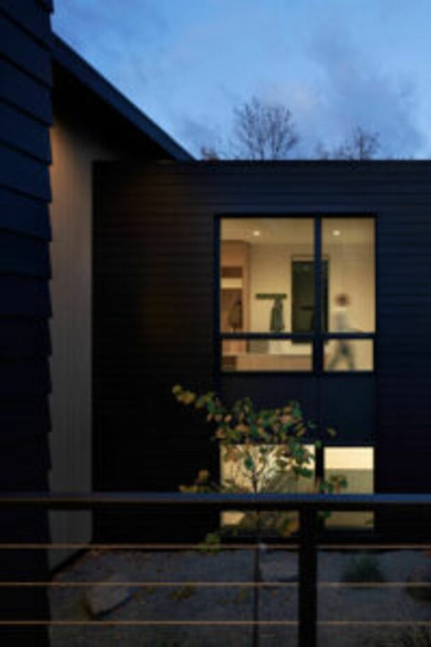 Twilight exterior view of a modern, compact building with dark horizontal cladding, a low-pitched roof, and large black-framed windows revealing a warmly lit interior. A small planted courtyard or balcony area and dark metal railing occupy the foreground.