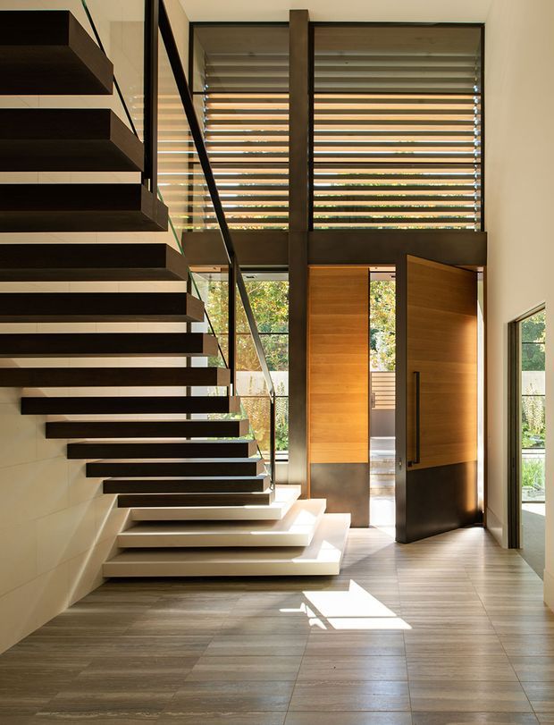 A bright, contemporary entry foyer featuring a dramatic floating staircase with dark timber treads and a slim glass-and-metal balustrade. Full-height glazing with horizontal timber louvres brings in filtered daylight and views of greenery. A tall timber pivot door opens to the exterior, while warm timber wall panels and large-format wood-look flooring create a refined, natural material palette.
