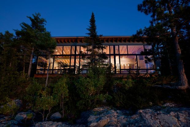 A modern glass-fronted house viewed from outside at dusk, nestled among trees and rocky natural terrain. Warm interior lighting illuminates the open-plan living spaces behind extensive floor-to-ceiling glazing, while a projecting timber roof structure and dark slender framing define the facade.