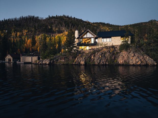 A lakeside house perched on a rocky shoreline, surrounded by dense forest in autumn. The building has a steep gabled section with exposed timber detailing, pale masonry or rendered walls, large glazed openings, and a dark pitched roof. A small waterside boathouse and dock are visible at the left, with the lake occupying the foreground.