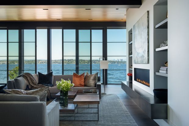 A refined contemporary living room overlooking a body of water through expansive floor-to-ceiling black-framed glazing. The room features a large sectional sofa, layered neutral and rust-toned cushions, a timber coffee table, grey rug, built-in dark shelving and cabinetry, a linear fireplace, and a warm timber-panelled ceiling. The space is bright, calm, and sophisticated, with a strong indoor-outdoor connection.