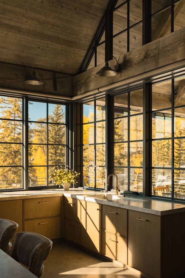 A warm, loft-like kitchen with extensive black-framed grid windows, timber cabinetry, and a pale stone benchtop. The room has a dramatic pitched timber ceiling and exposed heavy timber beams, with pendant lights above the work area. Strong afternoon sunlight enters through the glazing, overlooking autumn trees and an outdoor dining area.