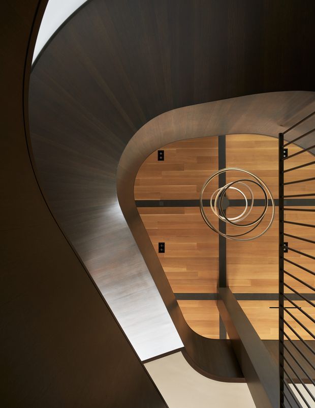 Dramatic architectural interior view looking down through a sculptural curved stairway or stairwell. Dark stained timber curves around a warm timber-clad vertical void, with black metal balustrade rails and a suspended concentric-ring light fitting. The composition emphasizes flowing geometry, layered levels, and strong contrast between dark wood, warm wood, and black metal.