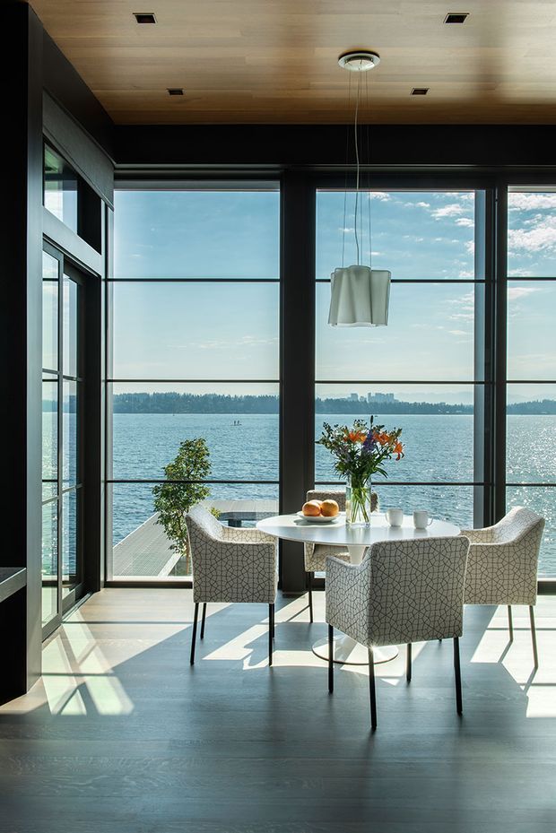 A bright contemporary dining area overlooking a body of water through expansive floor-to-ceiling black-framed glazing. The room features a round white dining table with four upholstered patterned chairs, a sculptural pendant light, dark timber flooring, and a light timber ceiling with recessed downlights. A small deck and distant shoreline are visible beyond the windows.