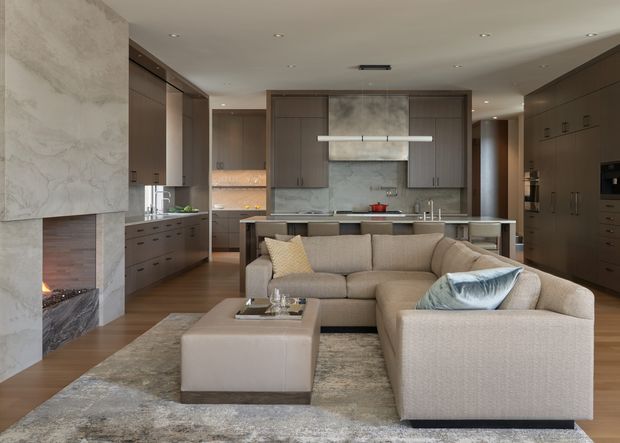 A refined open-plan interior combining a contemporary kitchen and living area. The room features extensive dark timber cabinetry, pale stone surfaces and splashbacks, integrated appliances, a central island, warm timber flooring, recessed ceiling lighting, and a large neutral sectional sofa arranged around a low upholstered ottoman. A stone-clad fireplace with visible fire anchors the left side of the space.