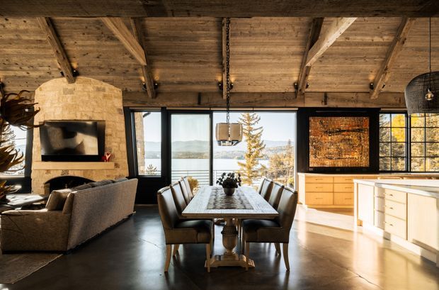 Warm open-plan interior combining a living room, dining area and kitchen. The space features a dramatic exposed timber-beam ceiling, dark polished concrete flooring, a rough light-stone fireplace surround, expansive black-framed glazing, a timber dining table with upholstered chairs, and light wood kitchen cabinetry with a large island. A lake and mountainous landscape are visible through the windows.