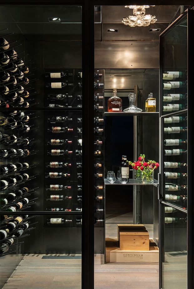 A dramatic, dark-toned wine storage room viewed through open glass-fronted cabinetry. Floor-to-ceiling wine racks display numerous bottles on both sides, while a mirrored central niche contains liquor bottles, decanters, wine glasses, flowers, and wooden wine crates. Black-framed glazing, reflective dark cabinetry, metallic wall panels, and a decorative crystal chandelier create a luxurious, gallery-like atmosphere.