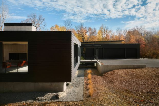 A low-slung modern residence with bold black horizontal cladding, flat rooflines, and large glazed openings. The house is set within a wooded landscape, with a concrete base, gravel paths, and a broad concrete terrace or driveway connecting to a dark enclosed volume. Warm-toned interior furnishings are visible through the glazing.