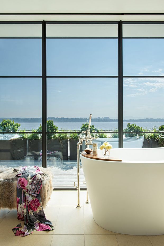 A bright, luxurious bathroom with a freestanding white bathtub positioned beside expansive black-framed floor-to-ceiling glazing. The windows overlook a body of water and distant shoreline, while potted greenery lines the exterior deck. A vintage-style brass bathtub tap and shower stand beside the tub, with a fur-covered stool and patterned textile adding warmth. The space has large pale neutral floor tiles and a serene waterfront atmosphere.