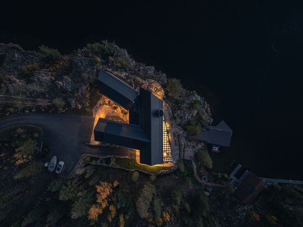 Aerial nighttime view of a modern angular residence set dramatically among rocky terrain and dense vegetation beside a dark body of water. The building has dark metal-clad roof and wall forms, warm exterior lighting, and a winding driveway with parked vehicles. A small waterside structure and jetty-like edge are visible to the right.