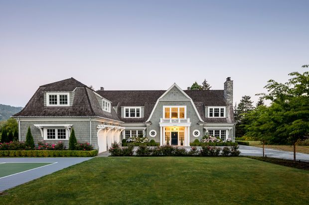 A large, symmetrical shingle-clad house viewed from the landscaped front lawn at dusk. The multi-part residence has several steep pitched and hipped roof sections, dormer windows, white-trimmed multi-pane windows, a prominent central gabled entrance with an illuminated upper balcony, and a tall stone chimney. Formal hedging, flower beds, paving, mature trees, and part of a tennis court surround the home.