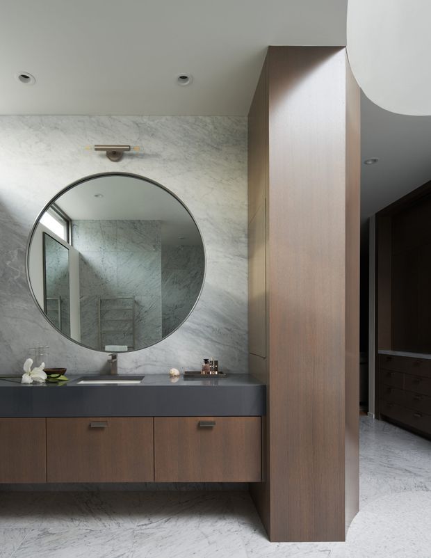 A refined contemporary bathroom vanity with a large round mirror mounted on a pale veined stone wall. The floating timber-toned cabinetry has a dark stone benchtop and integrated basin, with a brass-toned wall light above. Matching pale stone flooring and a tall timber storage partition create a calm, luxurious, minimalist interior.