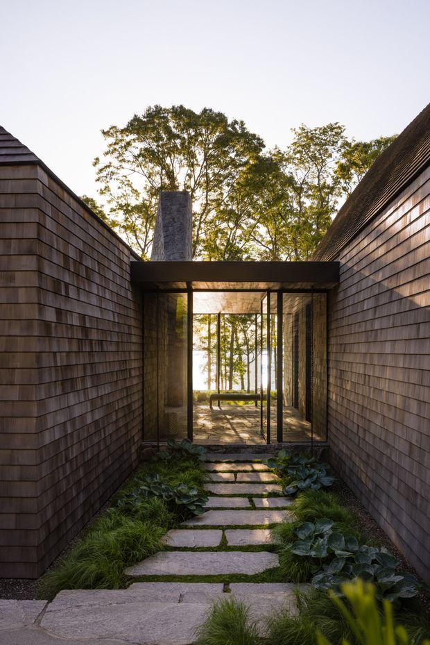 A narrow landscaped passage leads between two long shingle-clad building walls toward a glazed entrance or courtyard. Irregular stone stepping slabs are set among grasses and leafy planting, while a dark steel-framed glass opening reveals a sheltered seating area and mature trees beyond. A flat-roofed connecting volume and chimney-like masonry element create a strong framed architectural composition, with warm natural light and earthy textures.