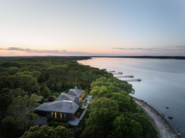 Aerial view of a dark-roofed, low-rise building nestled within dense coastal vegetation beside a calm waterway. A narrow shoreline and long timber jetties extend into the water, with a broad horizon and soft sunset light creating a secluded waterfront setting.
