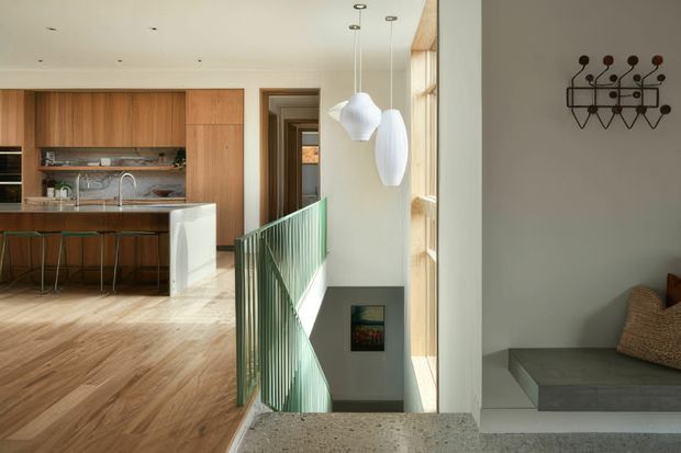 Bright contemporary open-plan interior showing a timber-lined kitchen with island and bar stools to the left, a stairwell with green metal balustrade in the centre, and a recessed grey built-in seating area to the right. Large windows bring in abundant natural light, while sculptural pendant lights and a geometric wall-mounted coat rack add character.