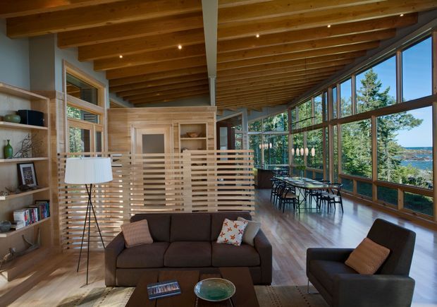 A warm, open-plan living room with a dark upholstered sofa and armchairs, a timber coffee table, built-in shelving, and a partially screened kitchen or dining area. Exposed timber beams and a sloping ceiling run toward extensive floor-to-ceiling glazing, which provides views of trees and water. The space combines natural timber finishes, recessed lighting, and a dining table positioned beside the windows.