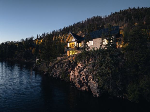 A warmly illuminated multi-level chalet-style house perched on a rocky forested lakeside or riverside cliff at dusk. The building has steep pitched gable roofs with dark roofing, light masonry or rendered walls, timber detailing, and large glowing windows and balconies. Dense coniferous woodland and a mountainous slope form the backdrop, with dark rippling water in the foreground.