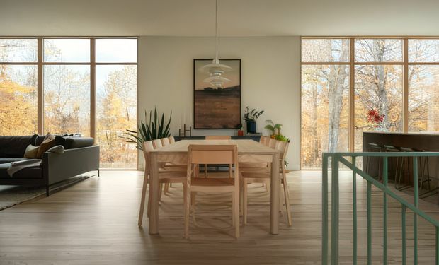 Open-plan dining area with a light timber dining table and chairs, large floor-to-ceiling timber-framed windows, pale wood flooring, and a living room visible to the left. A stair railing and kitchen or bar area appear on the right. The space is filled with warm natural light and overlooks autumn trees.