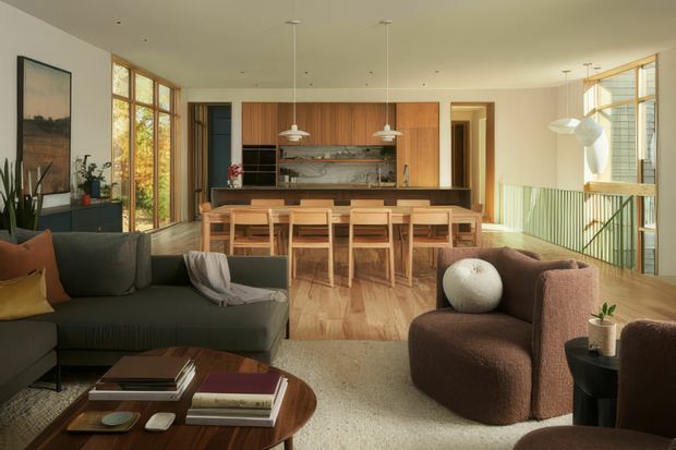 Open-plan contemporary living and dining space with a warm timber kitchen at the rear. A large dark sectional sofa and rounded brown armchairs frame a low timber coffee table in the foreground, while a long timber dining table sits beneath pendant lights. Floor-to-ceiling timber-framed glazing brings in abundant natural light, with a stair balustrade visible on the right and a softly textured neutral rug across the living area.