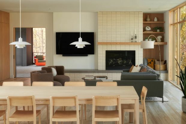 A warm, open-plan living and dining area with a large light-wood dining table and matching chairs in the foreground. Beyond it is a relaxed lounge with a sectional sofa, coffee table and wall-mounted television, centred around a tiled fireplace with timber shelving and built-in display joinery. Large timber-framed windows bring in abundant natural light, while simple white pendant lights and pale timber flooring create a calm, contemporary interior.