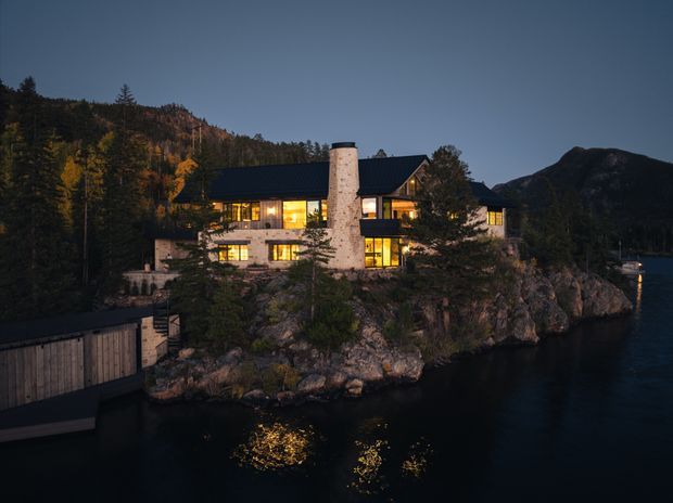 A warmly illuminated multi-level lodge-style house perched on a rocky waterfront site at dusk. The building has a dark pitched roof, extensive glowing windows, pale stone exterior walls, and a prominent cylindrical stone chimney. Dense autumn forest and mountainous terrain surround the property, with dark water directly below.