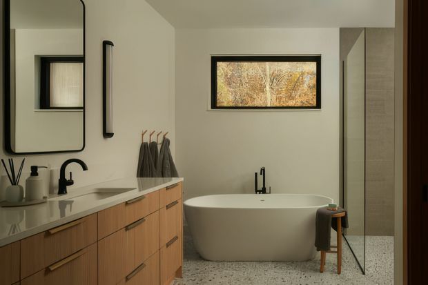 A warm, contemporary bathroom featuring a freestanding oval bathtub beneath a narrow horizontal window, a floating timber vanity with a pale stone benchtop, black tapware and hardware, and a large rounded-rectangle mirror. The room has light neutral walls, a terrazzo-style floor, and a glass-enclosed shower area at right.