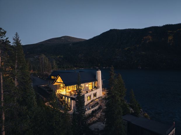 A dusk aerial view of a multi-storey lodge-style home set among dense forest beside a dark lake, with heavily wooded mountains rising behind it. The building has a steep dark pitched roof, warm illuminated glazing, a stone-clad lower level, and a prominent pale masonry chimney or vertical wall feature. A dark waterfront structure is visible in the lower right.