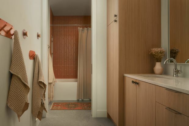 A warm, compact bathroom viewed from the vanity area toward a bathtub and shower. The room features timber-toned wall panelling and vanity cabinetry, a pale stone-look benchtop, white basin, chrome tapware, textured neutral towels on wall hooks, and a patterned bath mat. A timber-lined shower wall and light shower curtain create a cosy, retro-inspired interior.