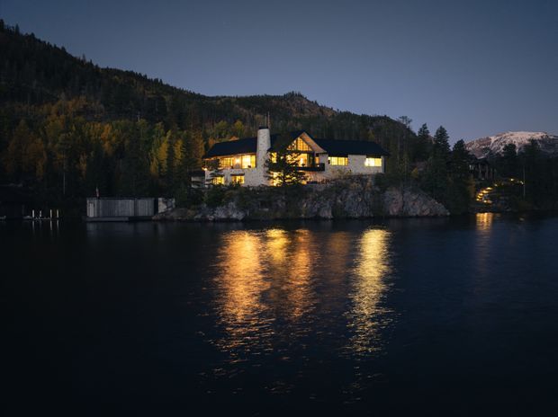 A warmly illuminated lodge-style house sits on a rocky, forested waterfront site at dusk. The dark pitched roof, prominent central gable, extensive timber detailing, and chimney are reflected in the calm lake, with wooded hills and distant snow-dusted mountains behind it.